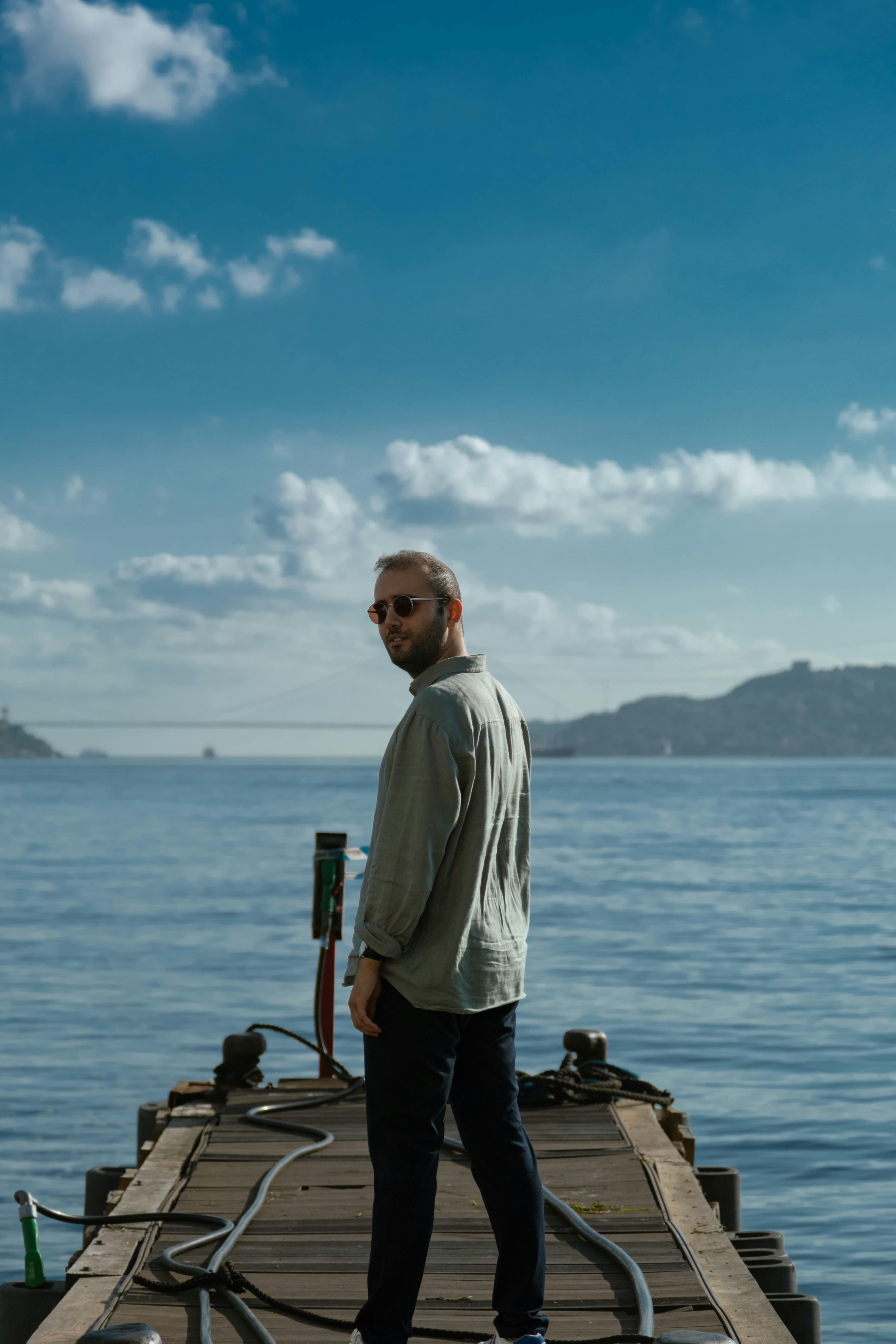 A man stands on a wooden pier gazing at the serene sea under a bright blue sky.