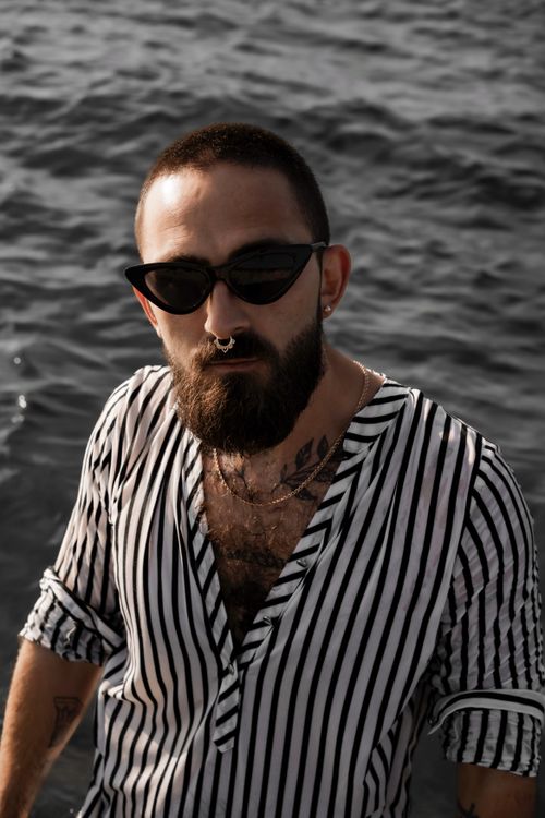 Free A stylish man with a beard and sunglasses standing by the sea, wearing a striped shirt. Stock Photo