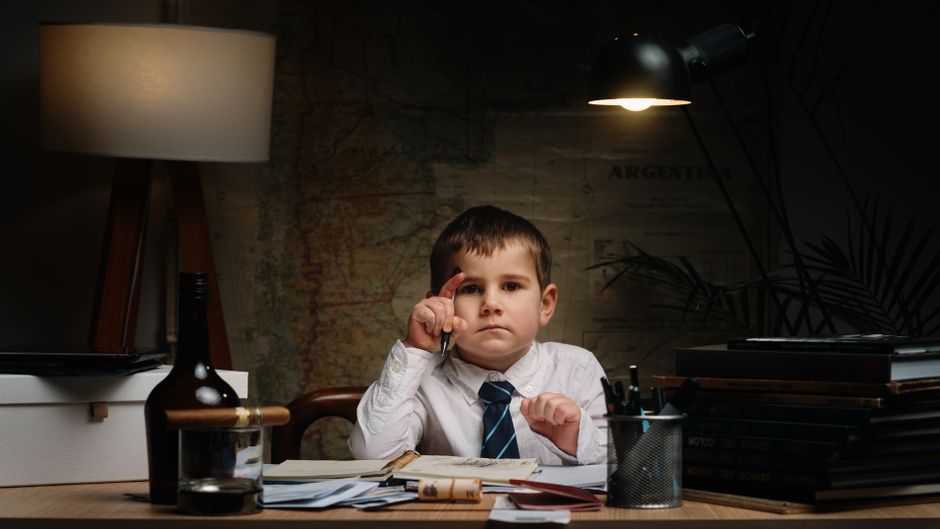 Un niño joven con camisa y corbata sentado en un escritorio con suministros de oficina, bajo una iluminación dramática.