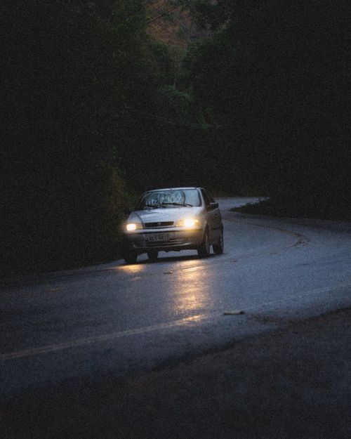 Free A car navigates a dark, wet road at dusk, headlights illuminating the way through a wooded area. Stock Photo