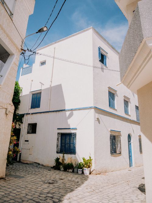 Free A beautifully sunlit Mediterranean-style white house with vivid blue accents on a quaint cobblestone street. Stock Photo