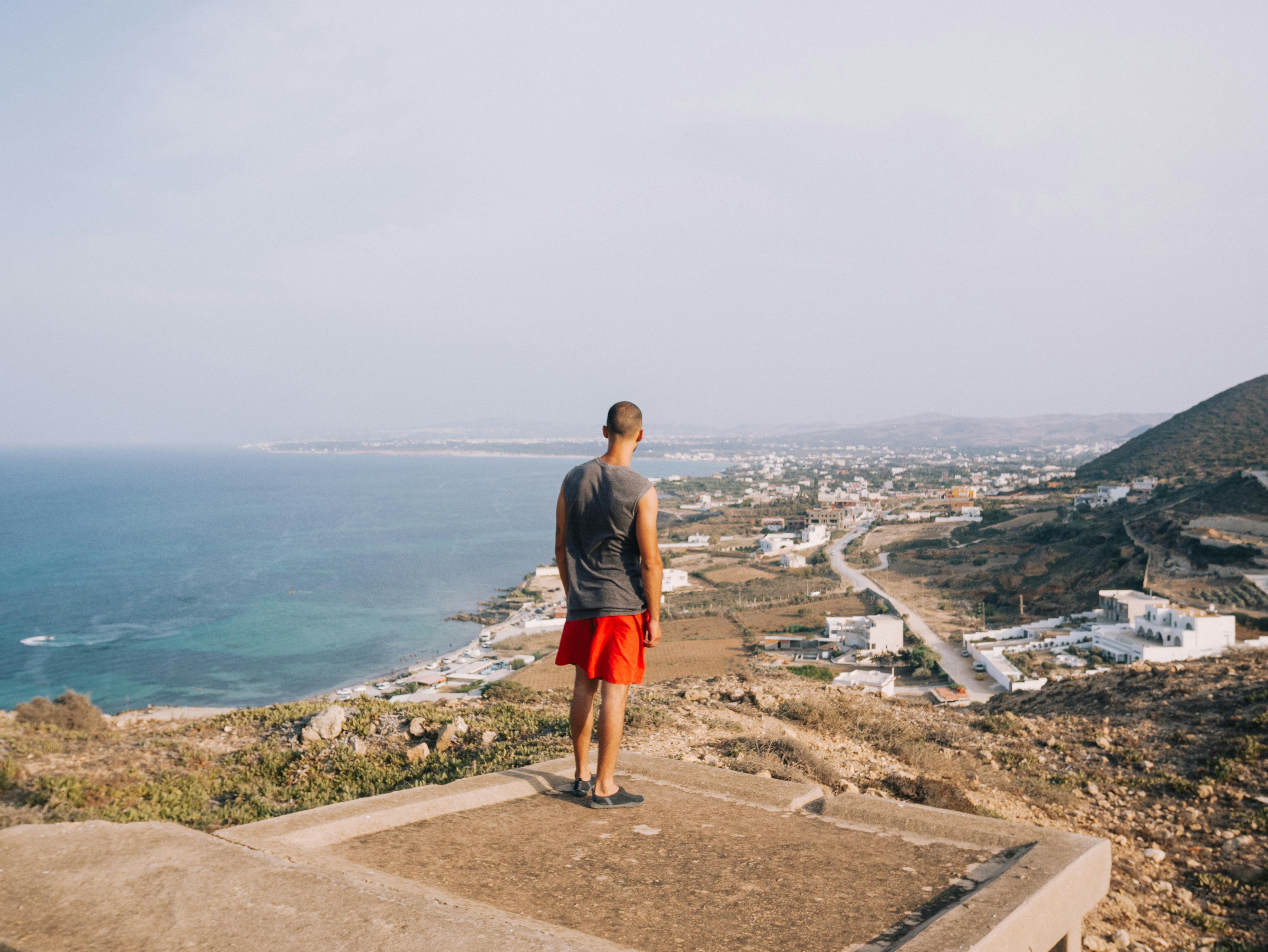 A man stands on a hill, overlooking a coastal cityscape, with the ocean extending into the distance.