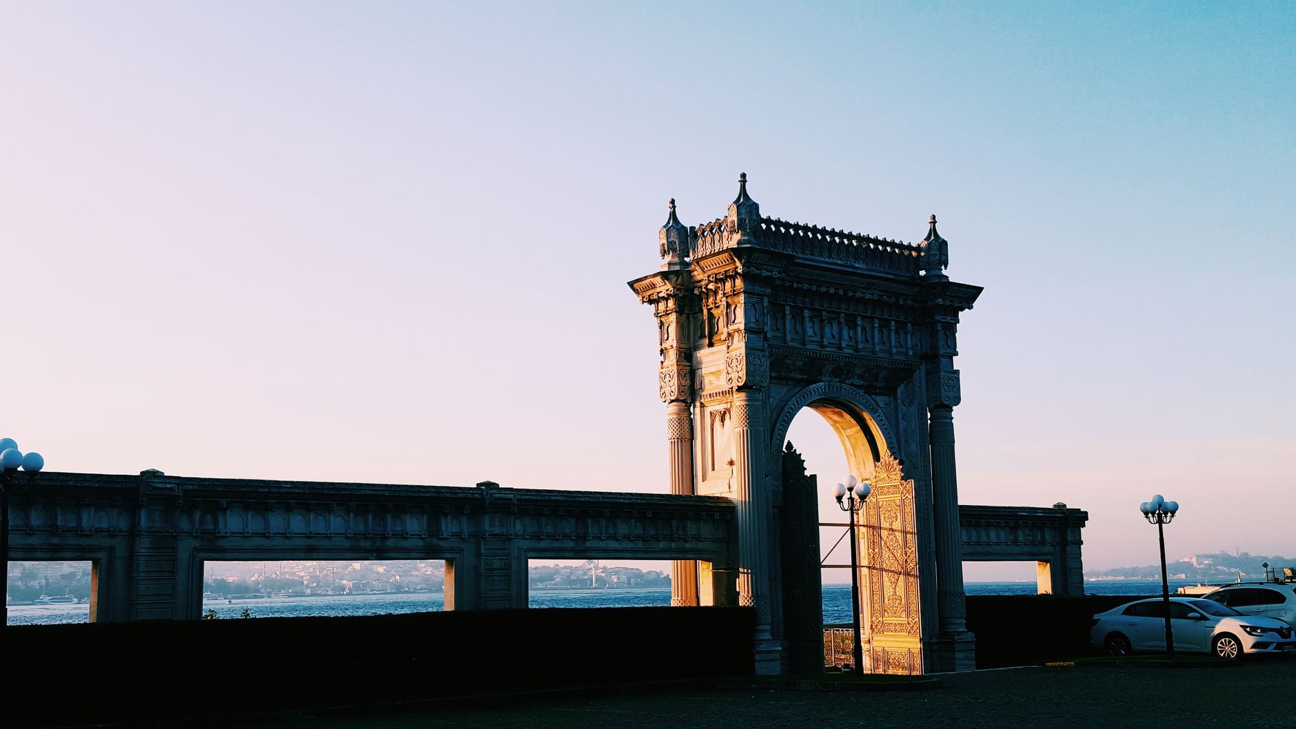 A beautifully lit historical gate by the Bosphorus in Istanbul during sunset with parked cars nearby.