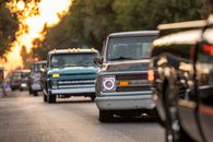 A lineup of classic vintage pick-up trucks cruising down a tree-lined road during sunset.