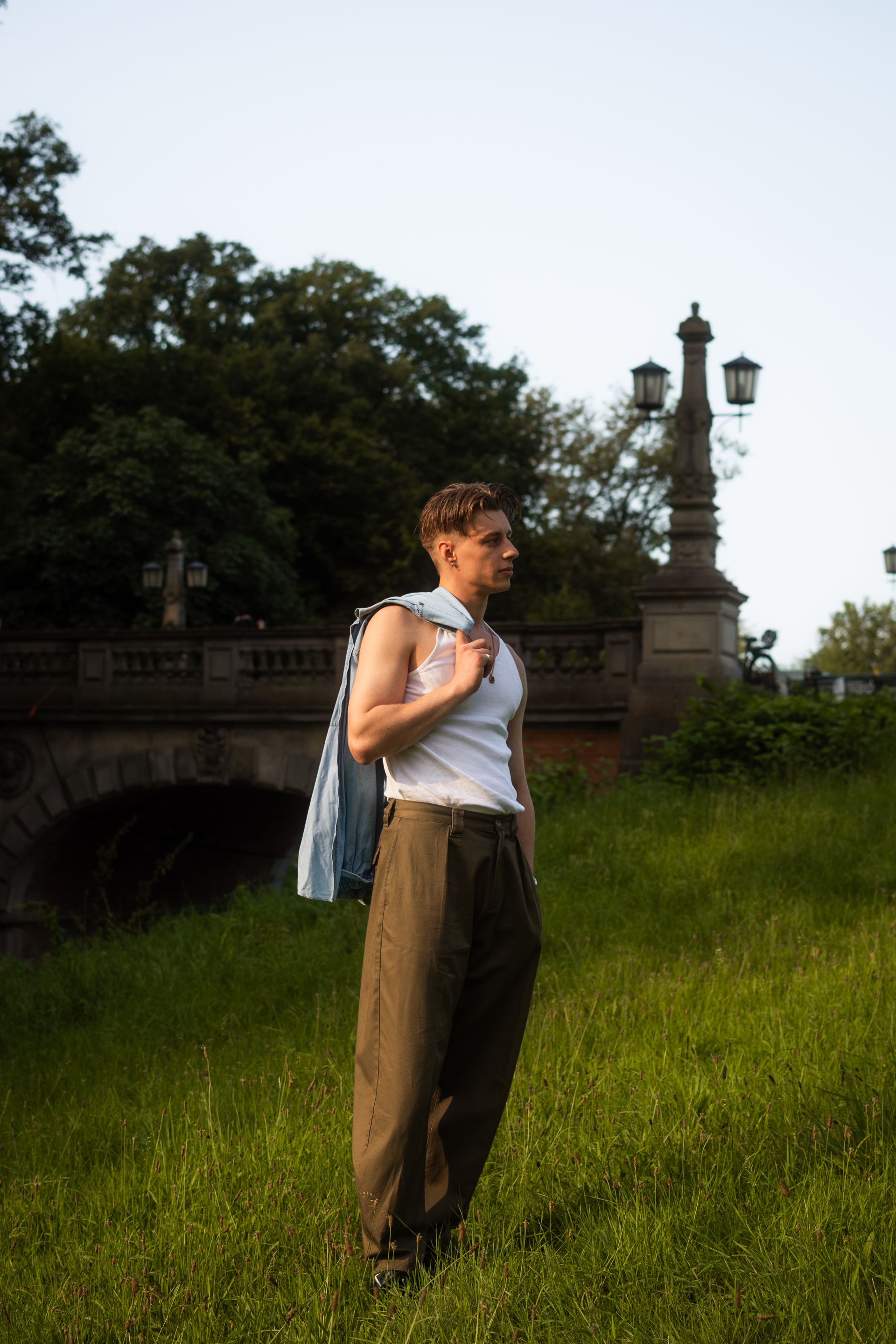A man in casual attire stands on a grassy meadow, basking in sunlight, with a scenic park bridge behind.