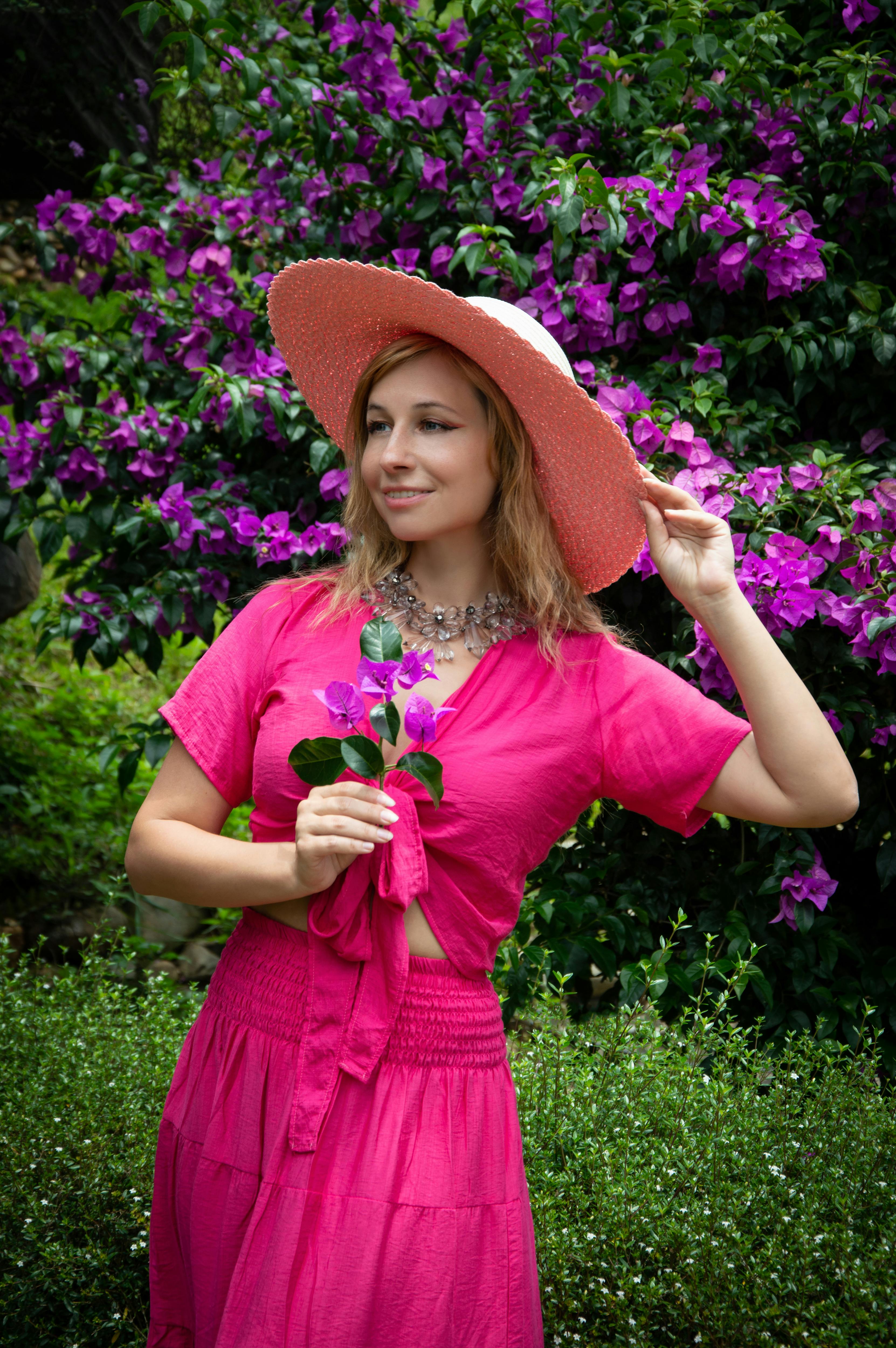 a woman in a pink dress and hat posing in front of purple flowers