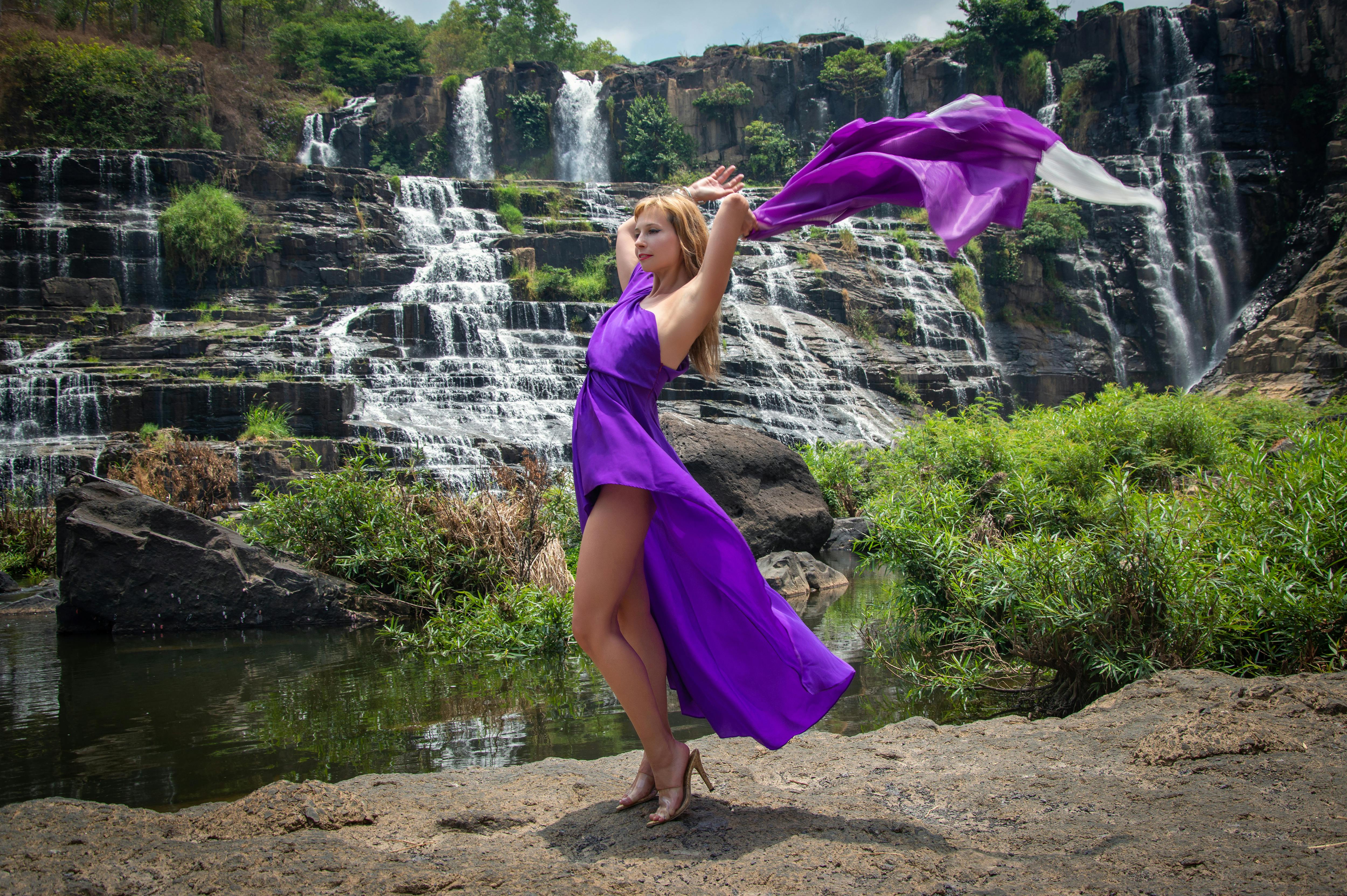 a woman in a purple dress is posing near a waterfall