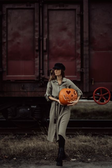 A woman carries a carved pumpkin with heart-shaped eyes, standing beside a train, embodying a Halloween vibe.