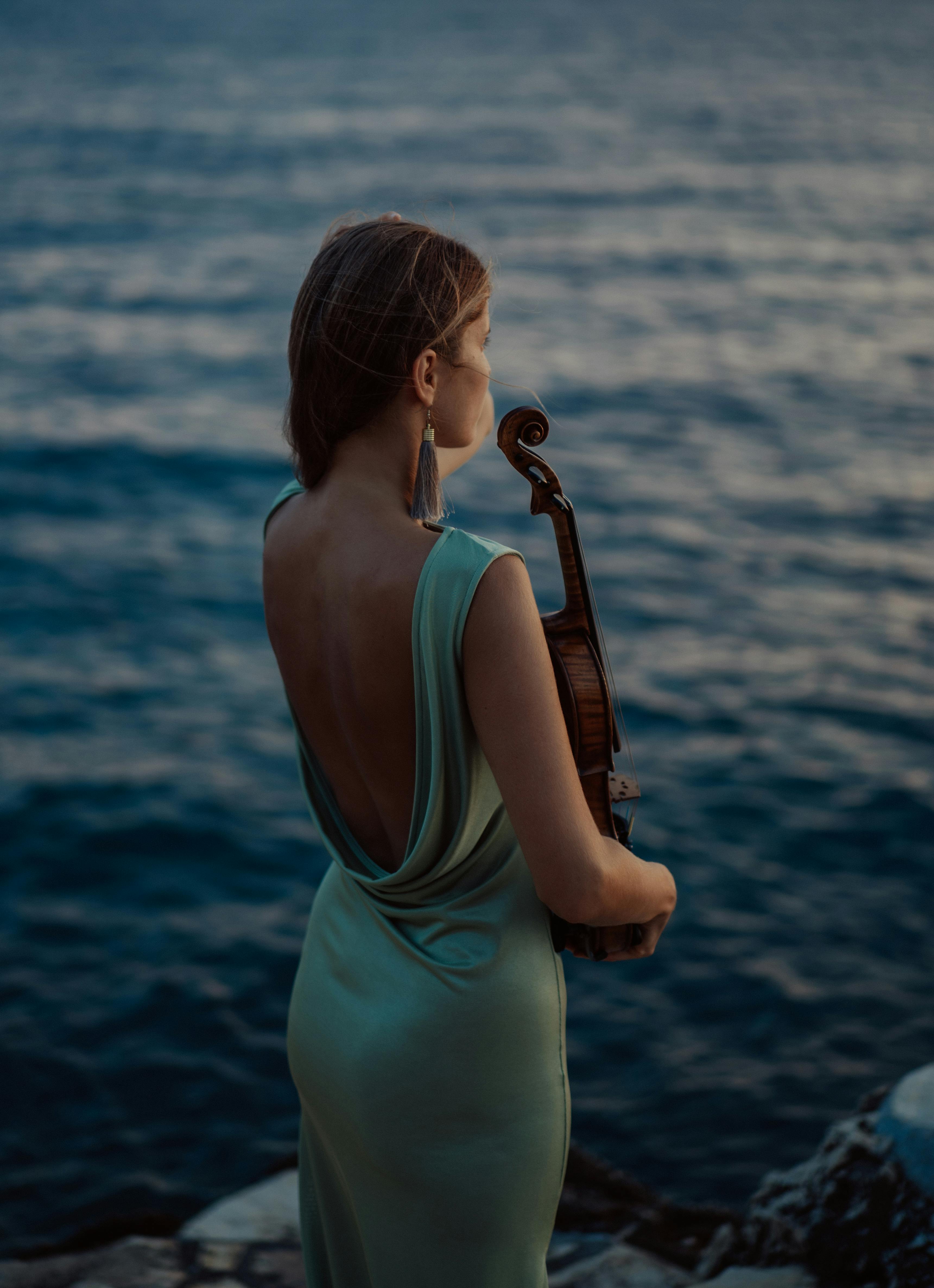 A woman in a green dress stands by the sea with a violin, captured at sunset in Rovinj, Croatia.