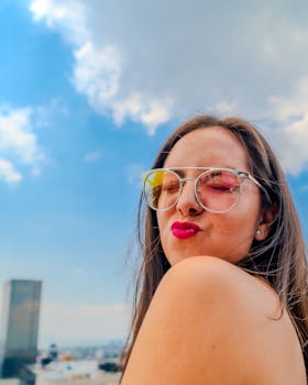 A cheerful young woman in sunglasses poses with a pout outdoors against a vibrant blue sky.