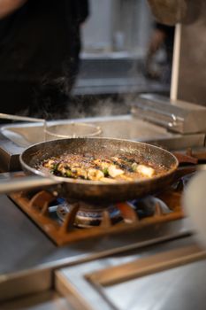 Sizzling fish dish prepared in a frying pan over a stove with visible flame and smoke in a modern kitchen.