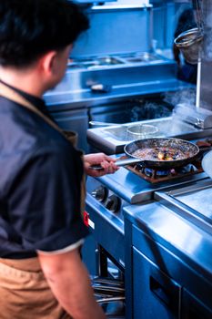 A chef in a black uniform cooking a dish in a pan on a stove in a professional kitchen.