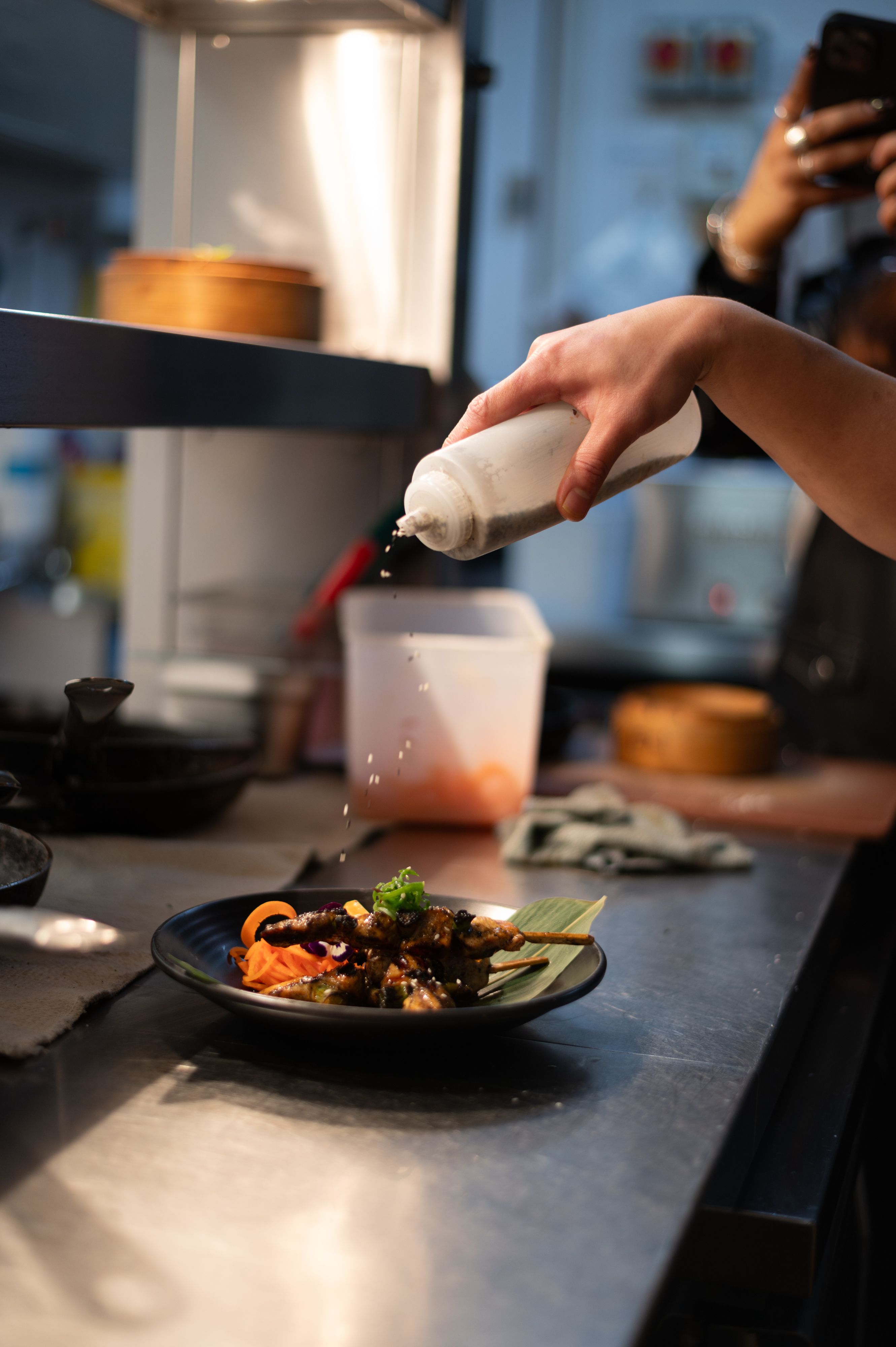 A hand garnishes a plated dish with sauce in a busy restaurant kitchen.