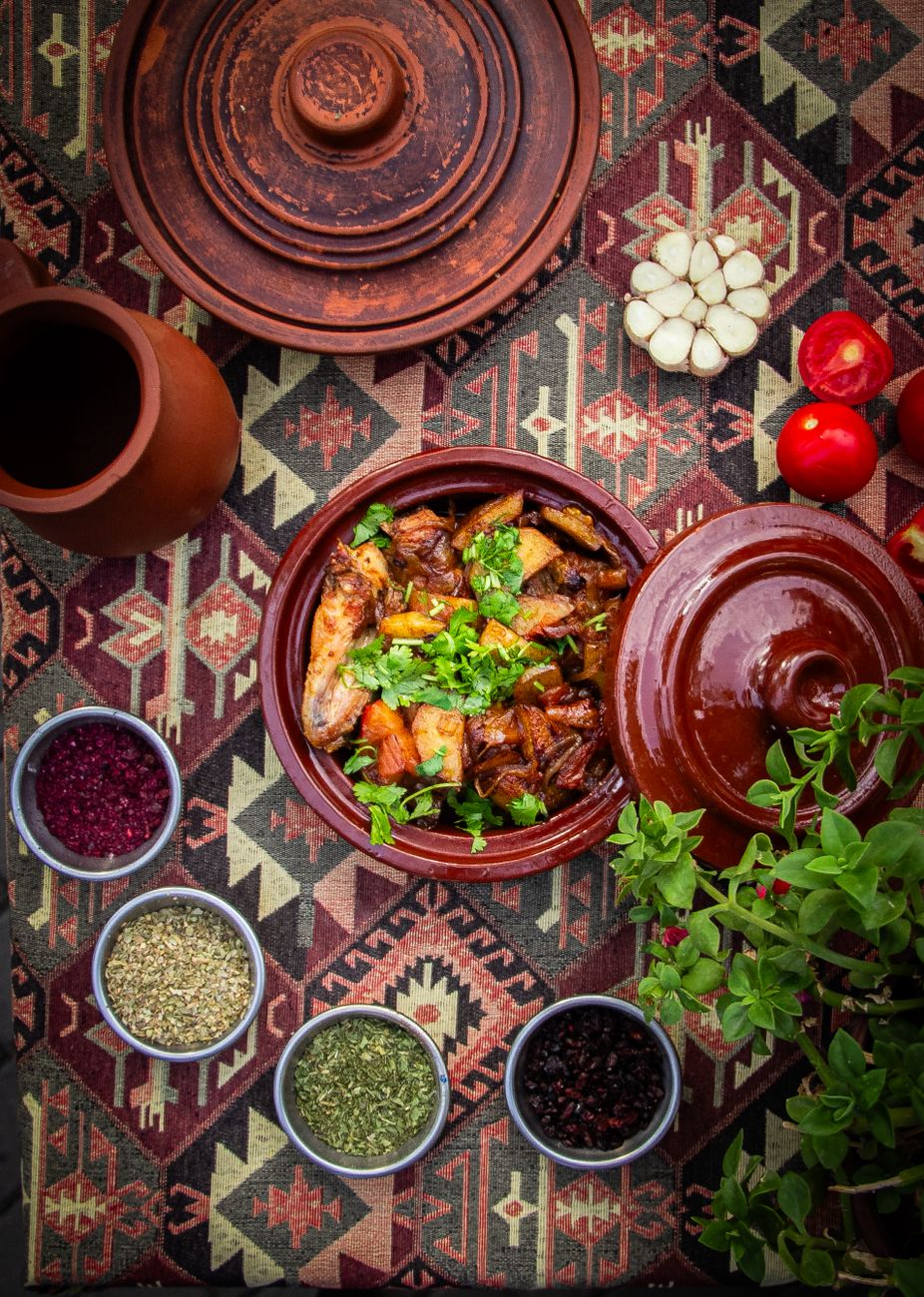 A traditional clay güveç pot filled with rich, mahogany-colored lamb stew, showing tender meat pieces, silky eggplant chunks, and whole red peppers, finished with a scatter of bright green parsley