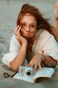 Woman enjoying a sunny beach day, relaxing with a book and camera.
