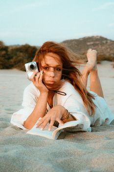 Woman lounging on sandy beach with camera and book, exuding summer relaxation.