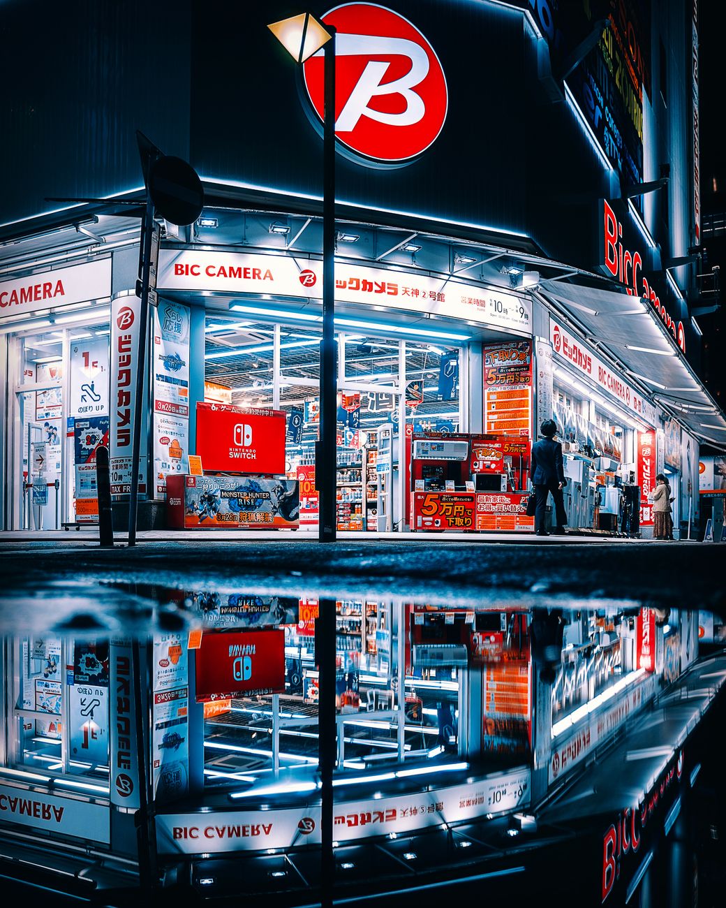 Nighttime scene of Bic Camera store with neon lights reflecting on wet street in Fukuoka, Japan.