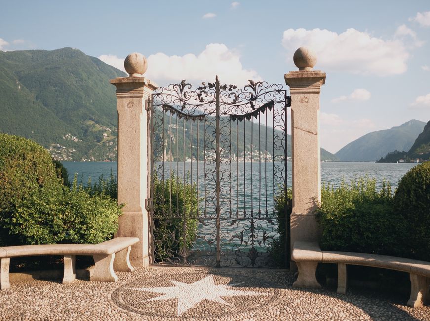An ornate iron gate with mountain and lake view, set in a serene garden in Bergamo, Italy.