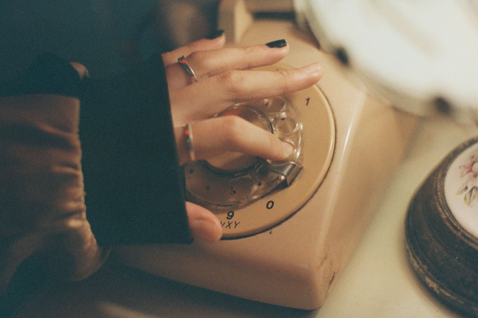A hand with dark nail polish dialling a cream coloured rotary telephone on a table, fingers in the dial and the cord curling away out of frame