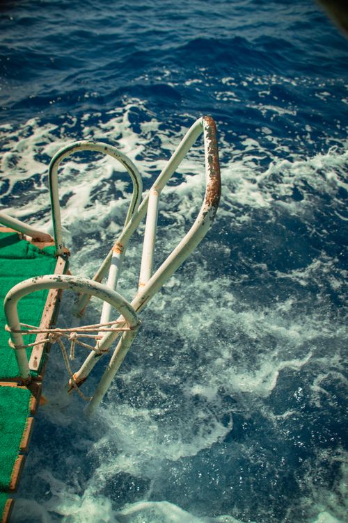 Free Rusty metal ladder descends into vibrant blue ocean water, surrounded by foamy waves. Stock Photo