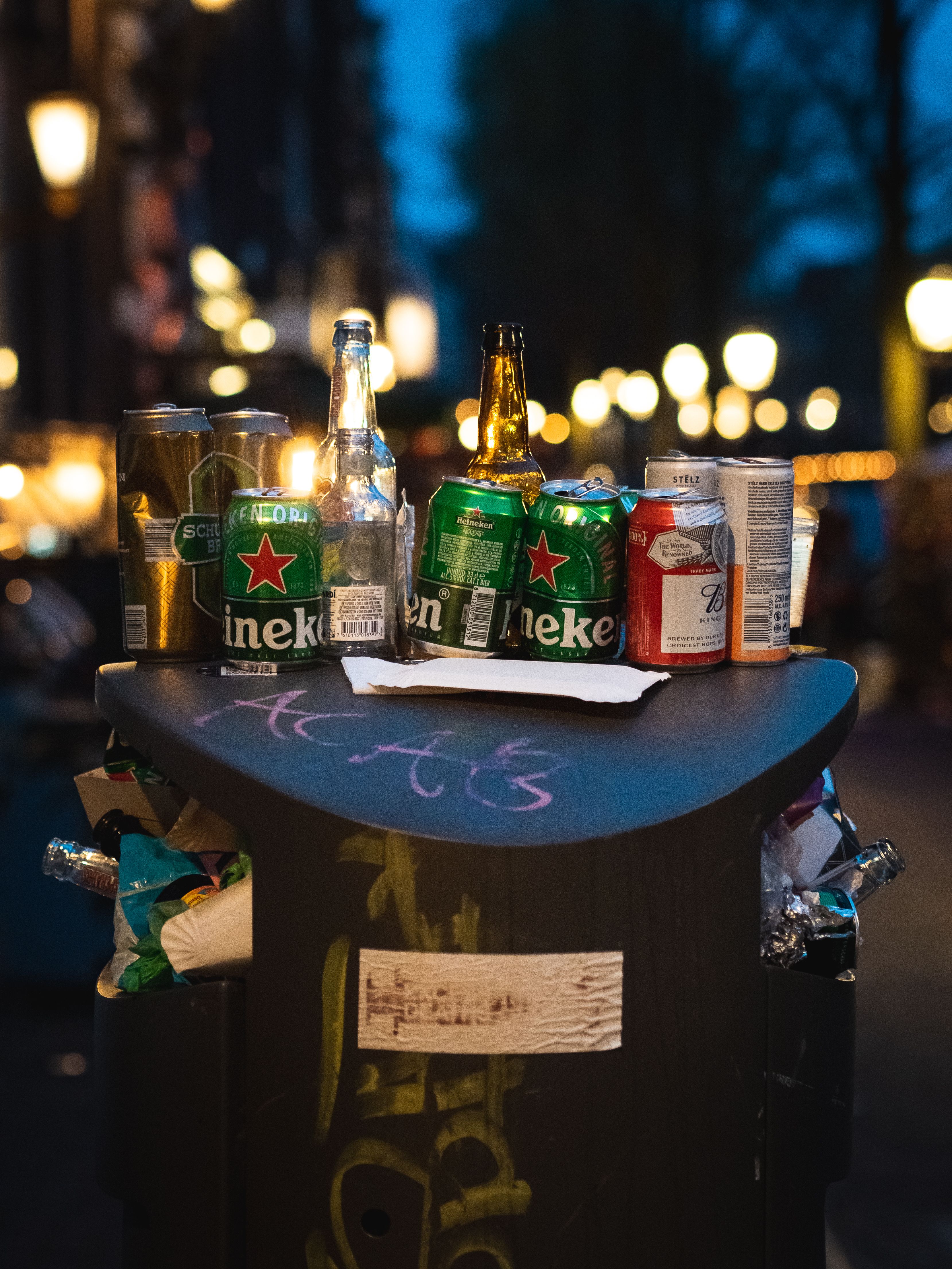 A vibrant street scene depicting discarded bottles and cans on a city trash can at night.