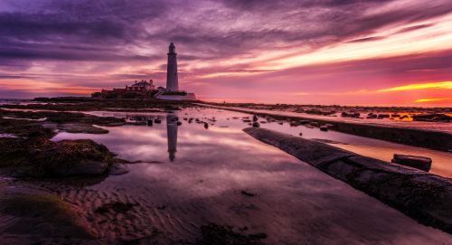Free Stunning sunset view of a tall lighthouse reflecting in calm tide pools on a rocky seashore. Stock Photo