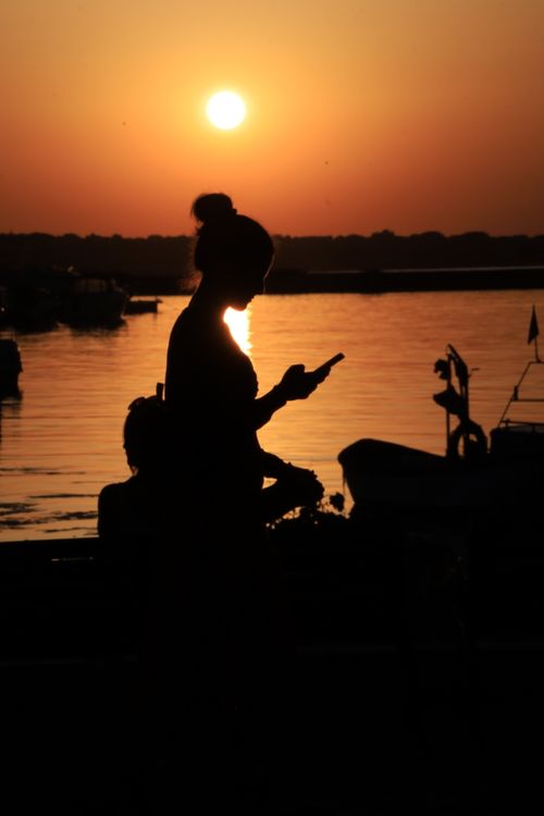 Free A woman checks her phone in silhouette against a vibrant sunset by the water, creating a serene scene. Stock Photo