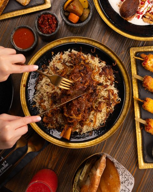 Free Close-up of hands cutting lamb served with rice, surrounded by appetizers and sides on a wooden table. Stock Photo