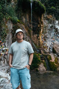 A young man stands confidently by a serene waterfall in Гагра, surrounded by lush greenery.