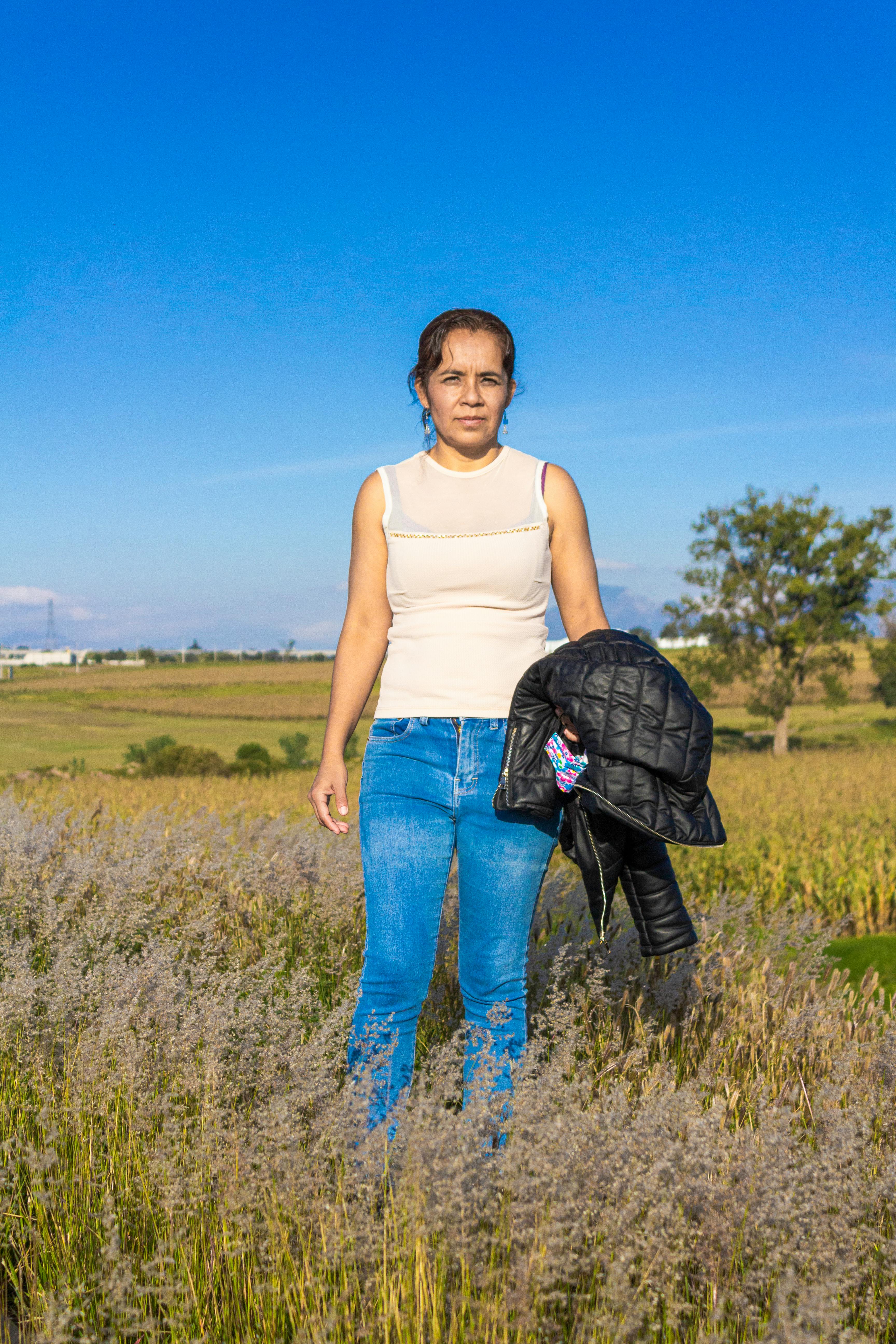 Woman in white top and jeans standing in a sunny field with a leather jacket.