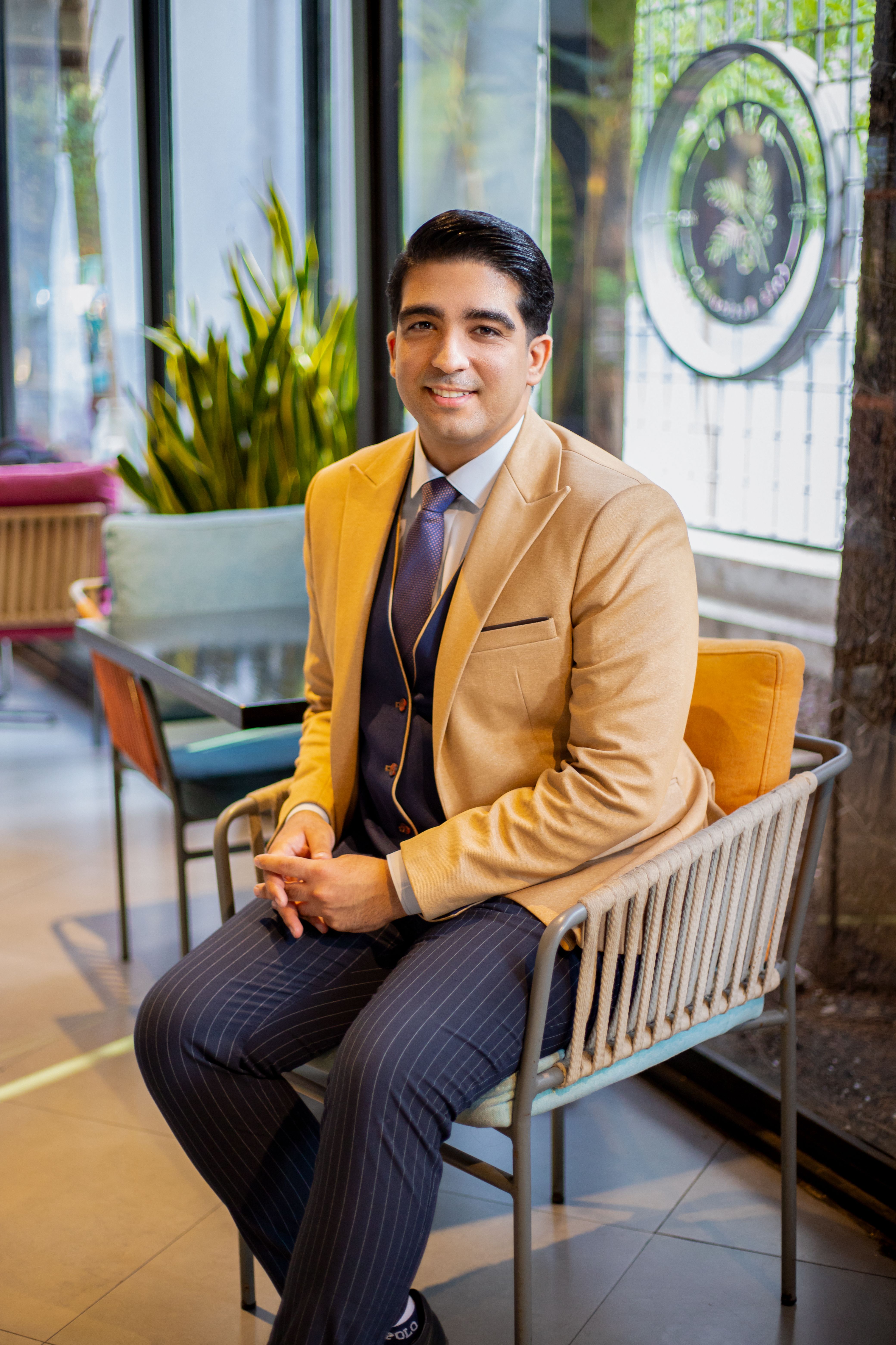 A young man in a beige suit smiling while seated in a contemporary café setting with lush greenery.