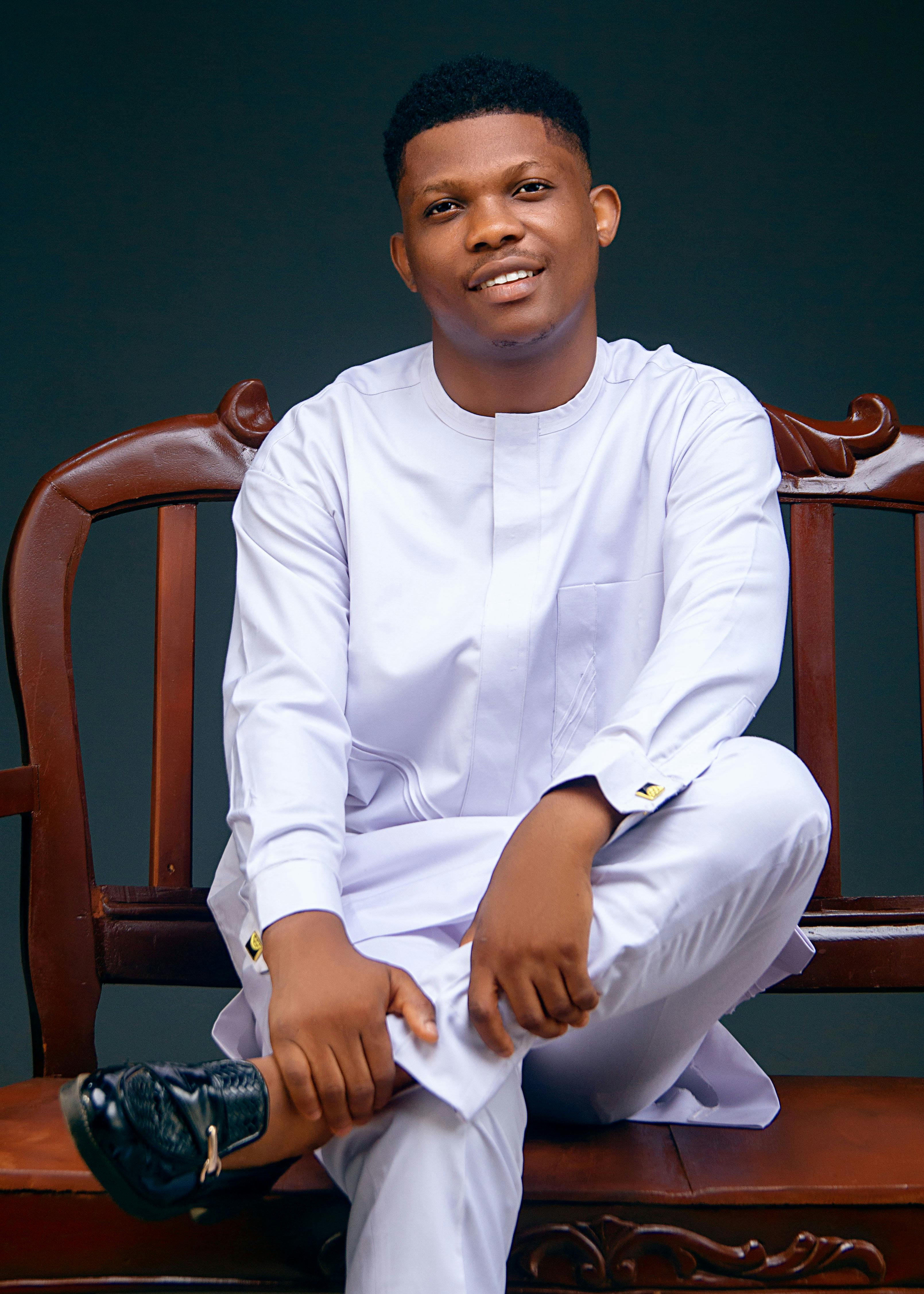 Portrait of a smiling young man in a white traditional outfit sitting on a vintage wooden bench indoors.