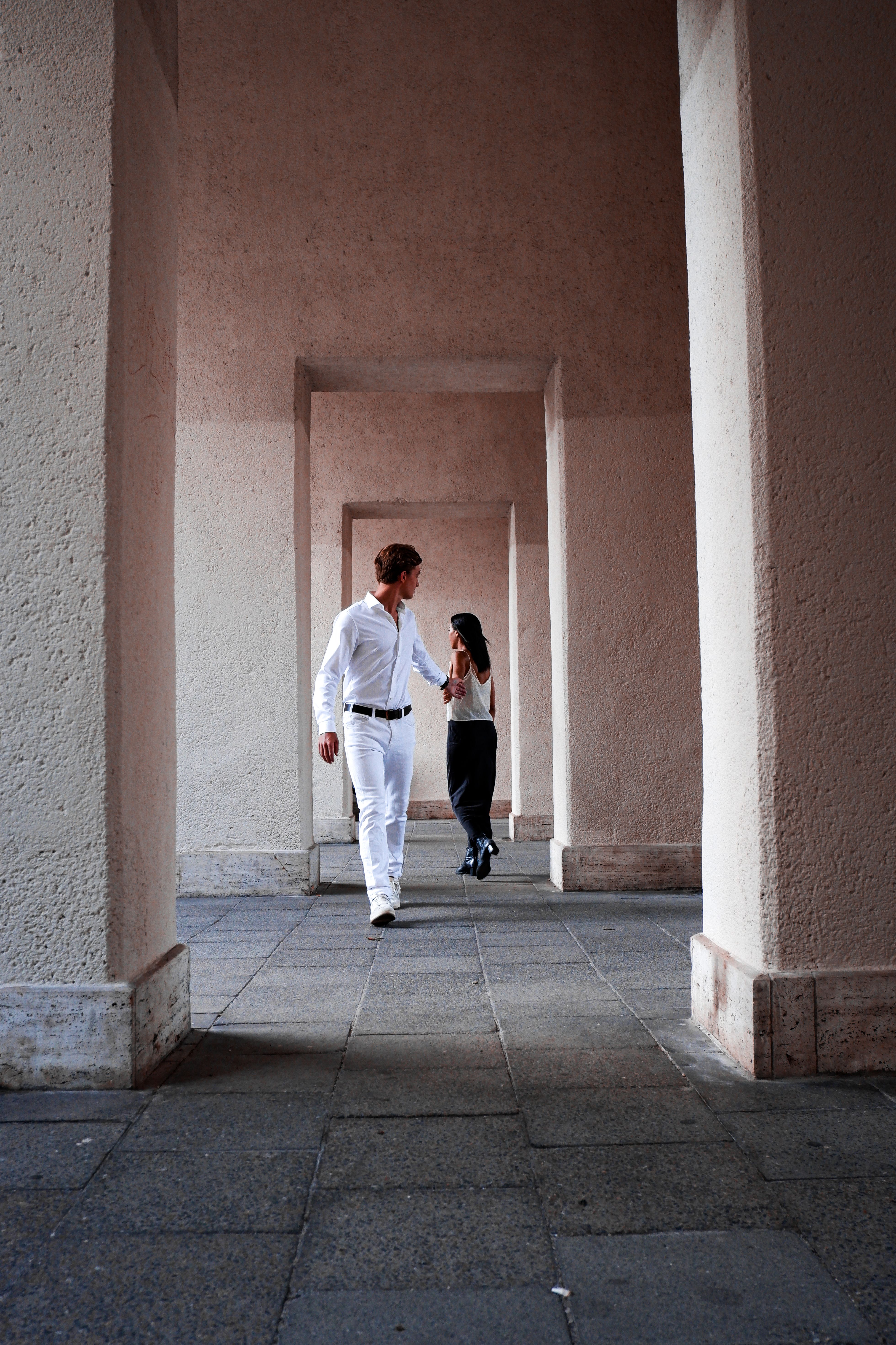 A stylish couple strolls through a dramatic architectural hallway filled with shadows and light.