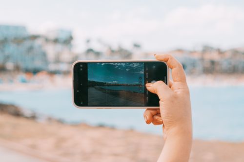 Free A person photographs a scenic coastal view using a smartphone against the backdrop of a beach. Stock Photo