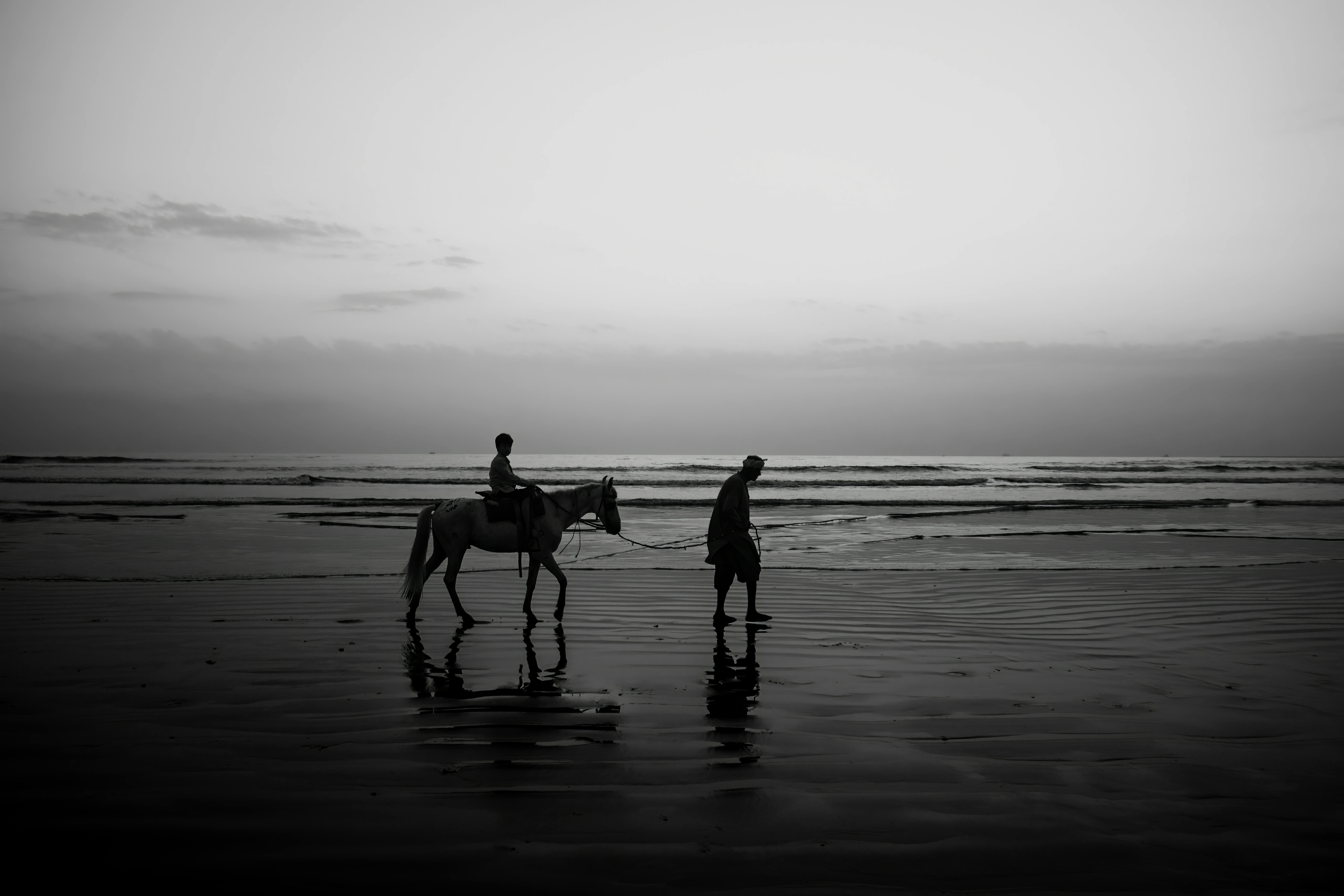 A serene black and white silhouette of a person riding a horse on Karachi beach during sunset, capturing a tranquil coastal moment.
