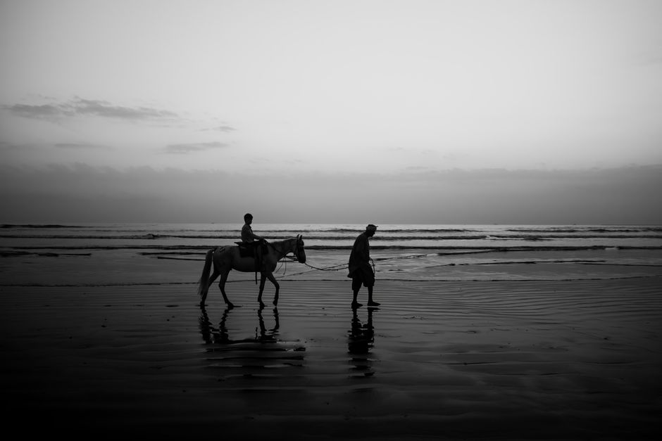 A serene black and white silhouette of a person riding a horse on Karachi beach during sunset, capturing a tranquil coastal moment.