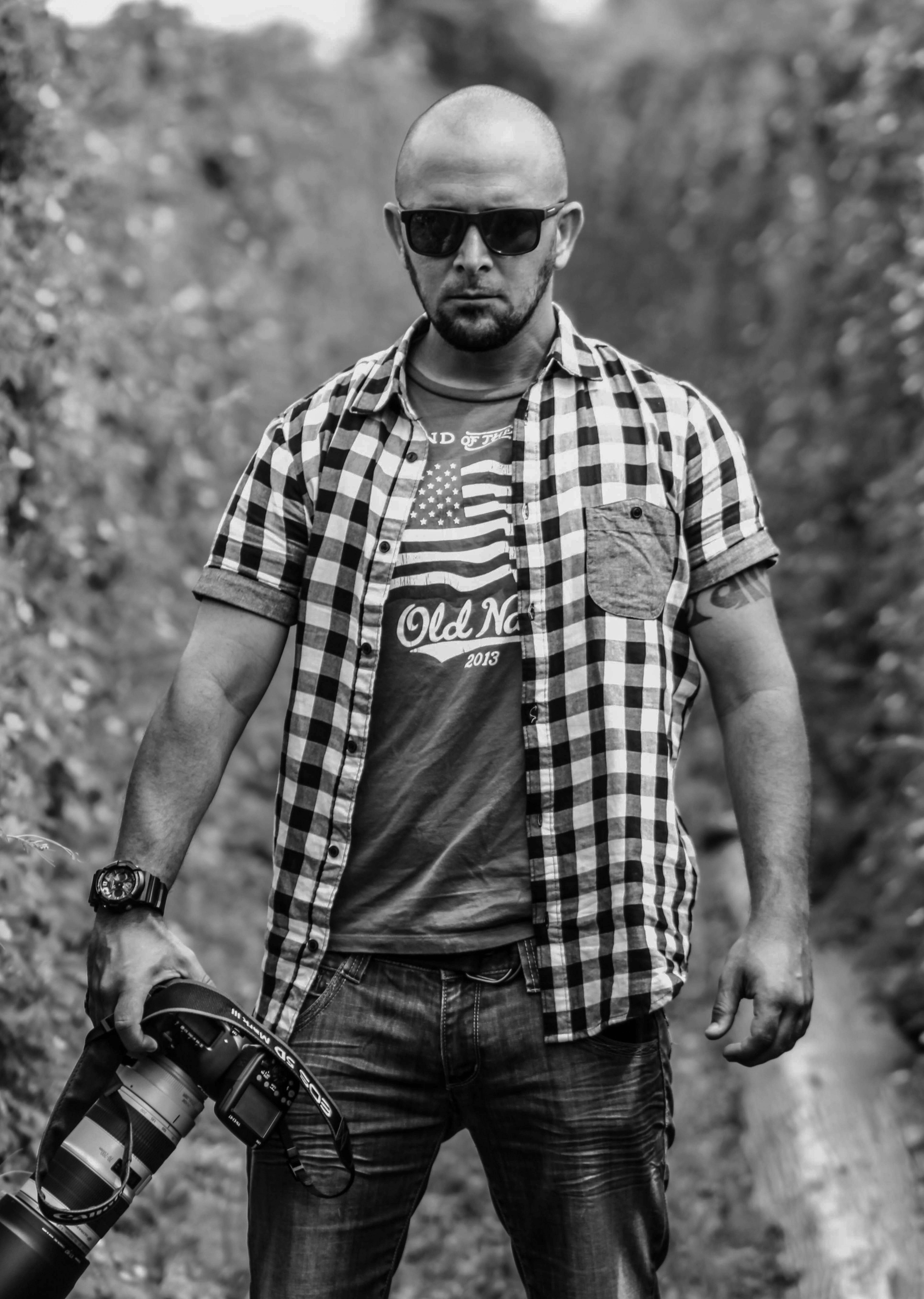 Black and white portrait of a man in plaid shirt, holding camera, outdoors.