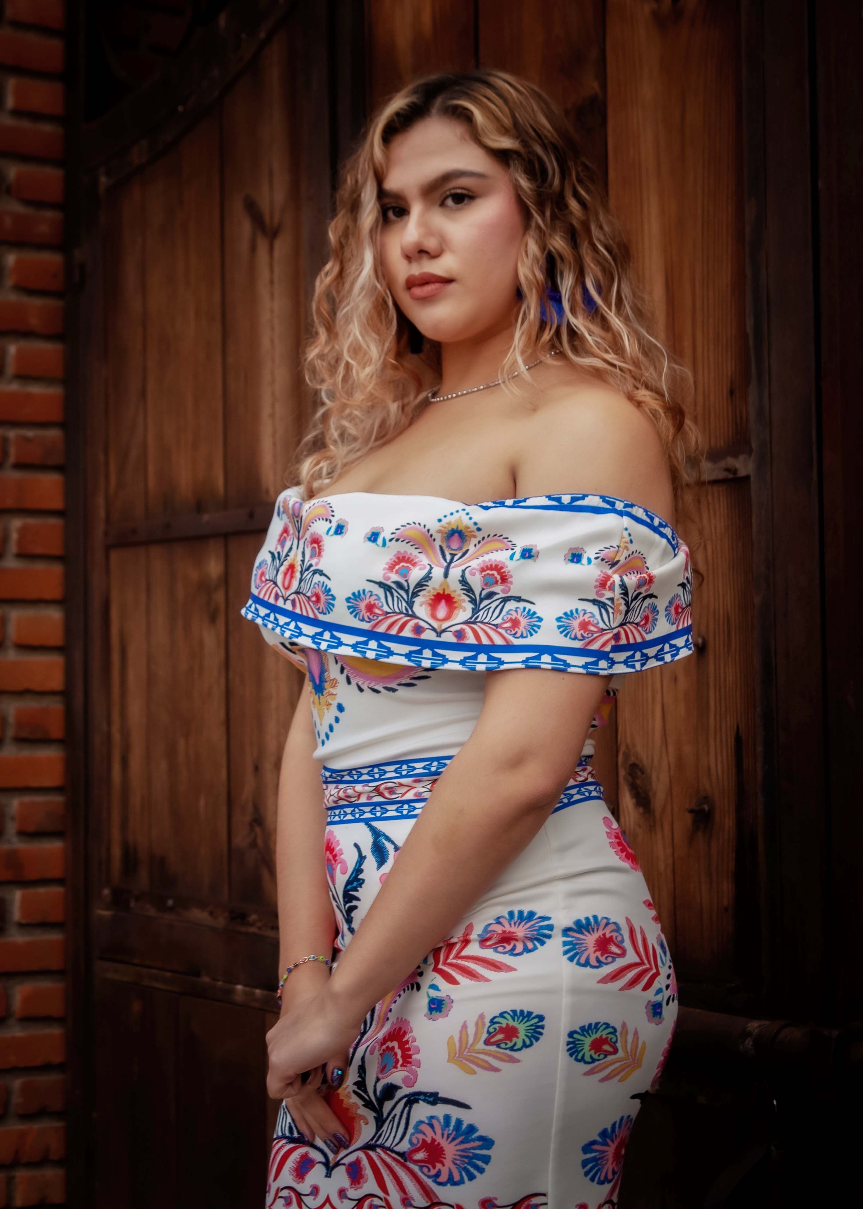 Portrait of a young woman in a floral dress standing elegantly in front of a rustic wooden door.