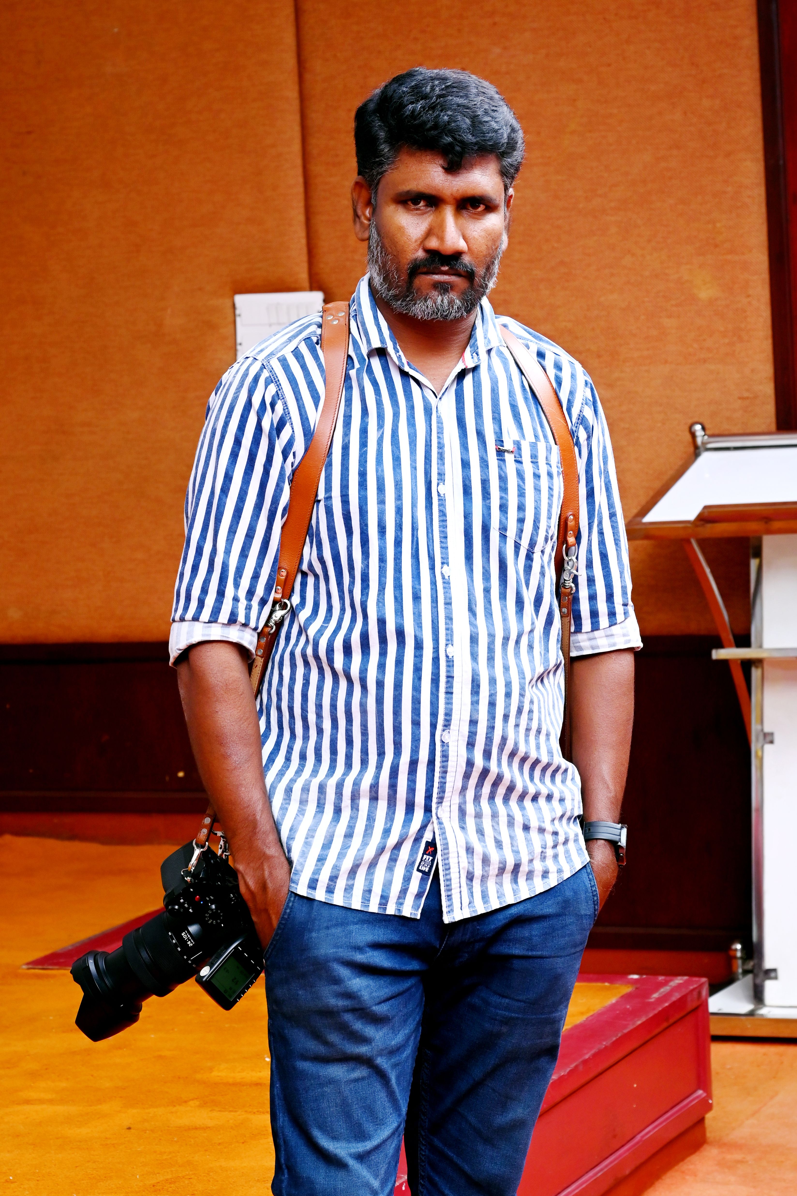 A photographer poses indoors with camera gear, wearing a striped shirt and jeans.