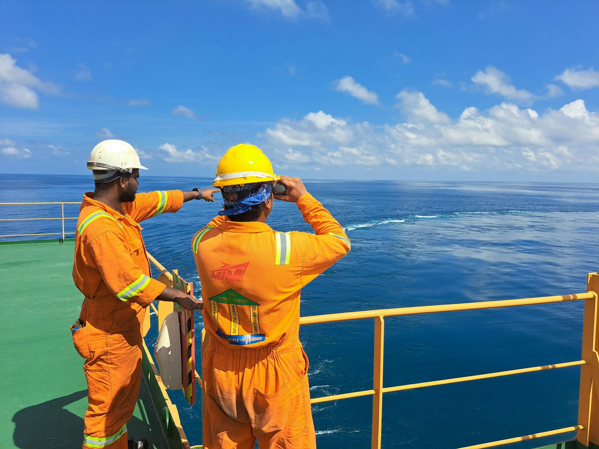 Two workers on an oil rig deck wearing safety gear inspect the ocean horizon under a clear sky.