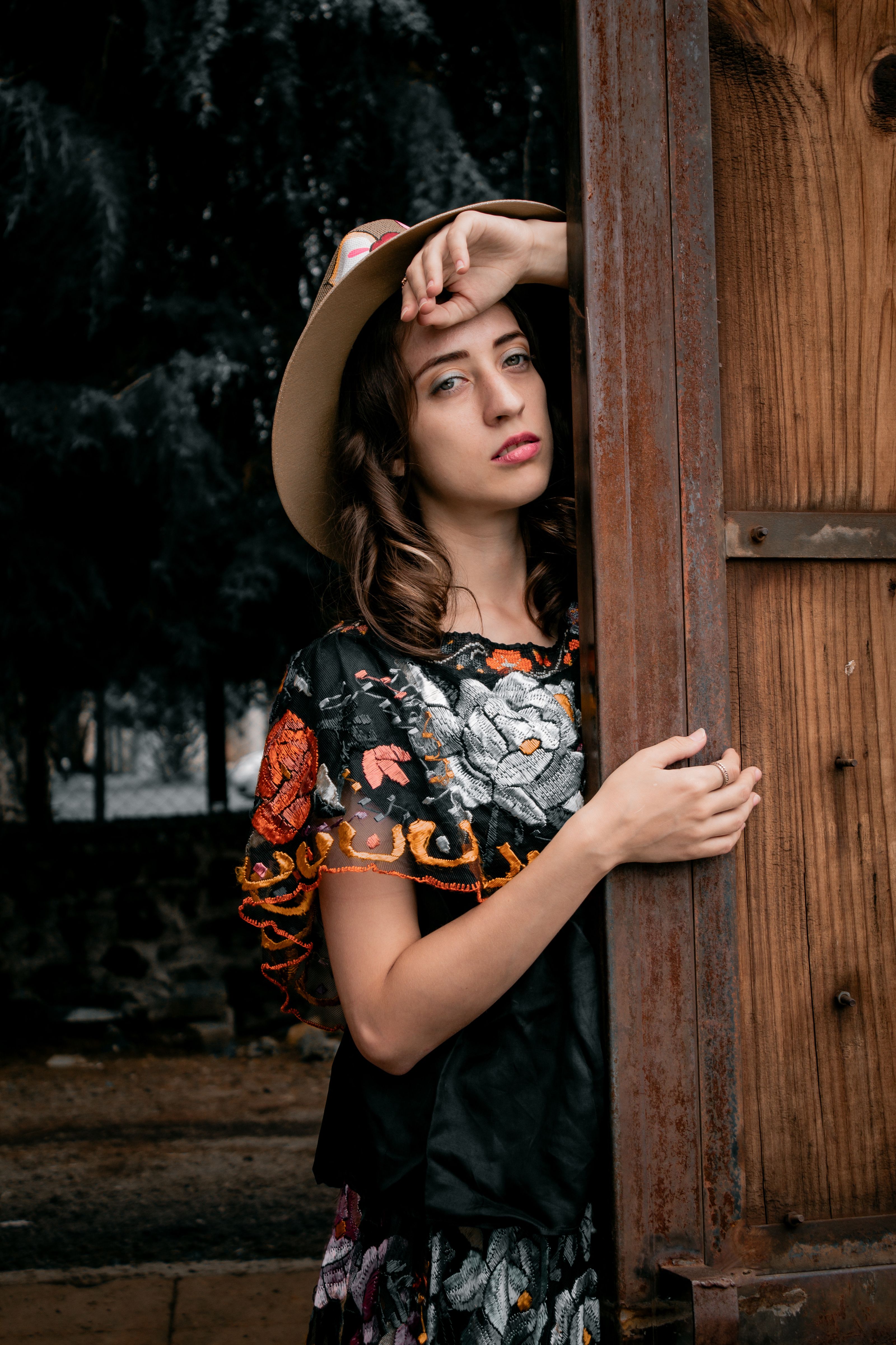 Uma jovem mulher em um vestido bordado floral e chapéu posa ao ar livre, apoiando-se em uma porta.