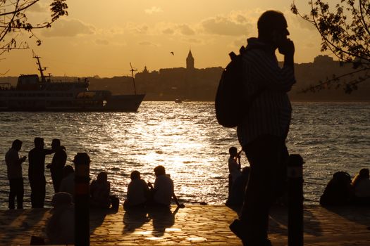 A scenic view of people silhouetted against the sunset by the water with a cityscape in the background.