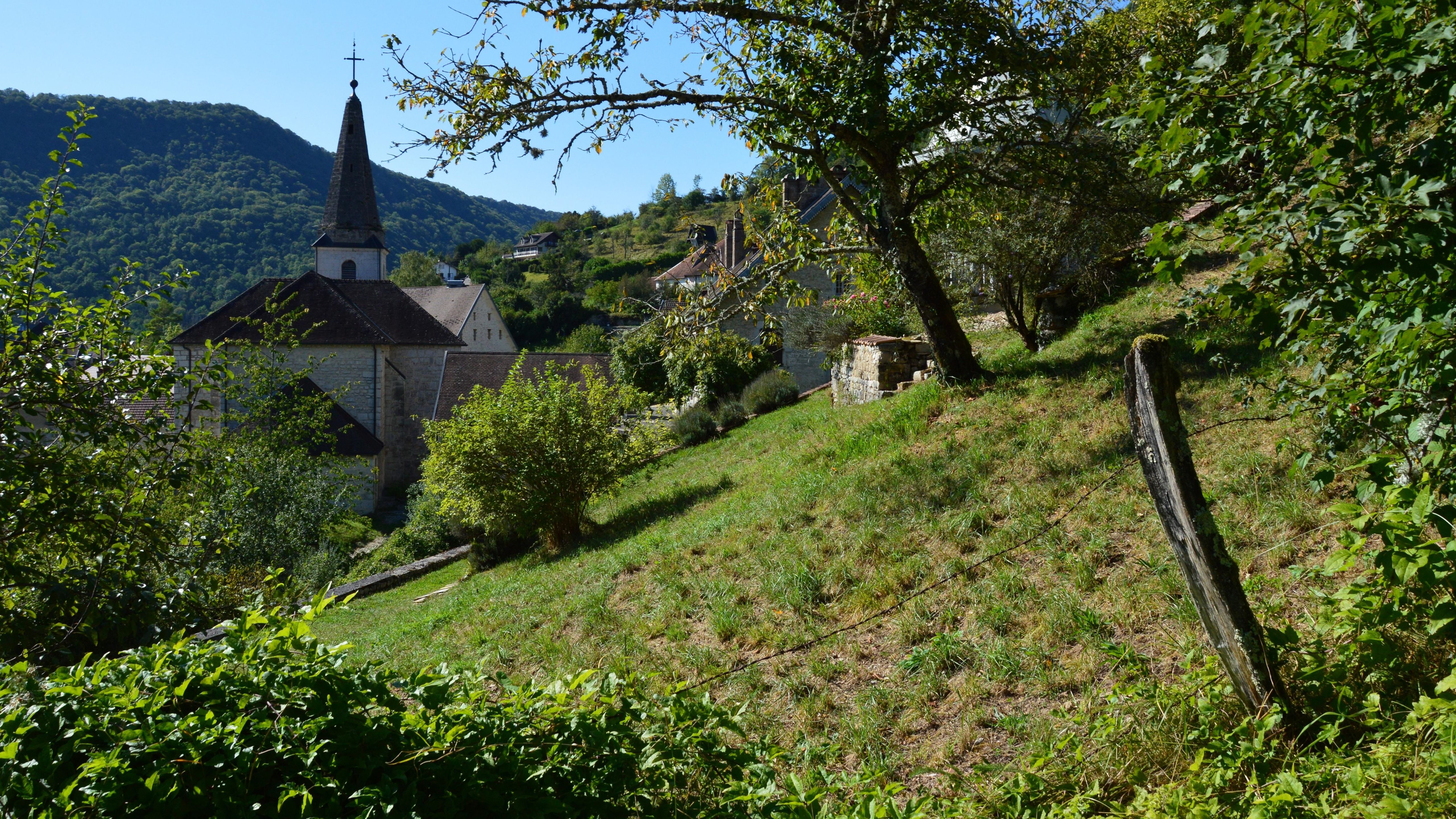 Sonniges Hanggrundstück mit Wiese am Ortsrand eines europäischen Dorfes mit Kirchturm und bewaldeten Hügeln im Hintergrund