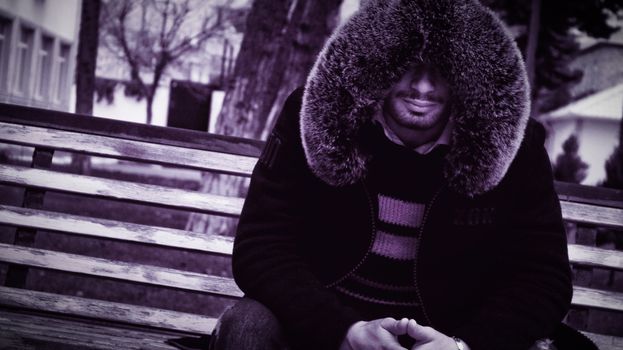 A mysterious man in a fur hooded coat sits on a park bench exuding a cool vibe.