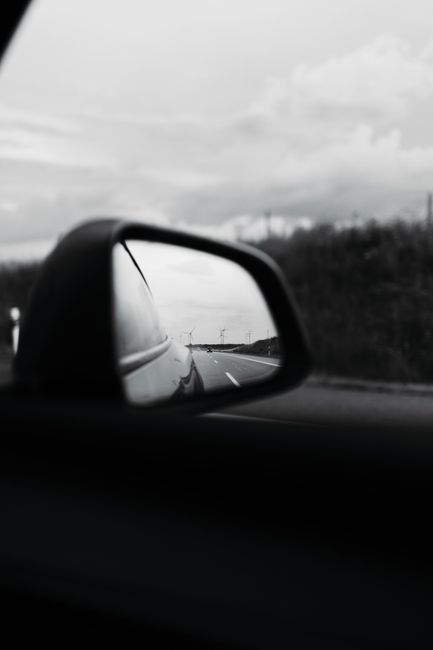 A black and white photo capturing a road reflected in a car mirror on a cloudy day.