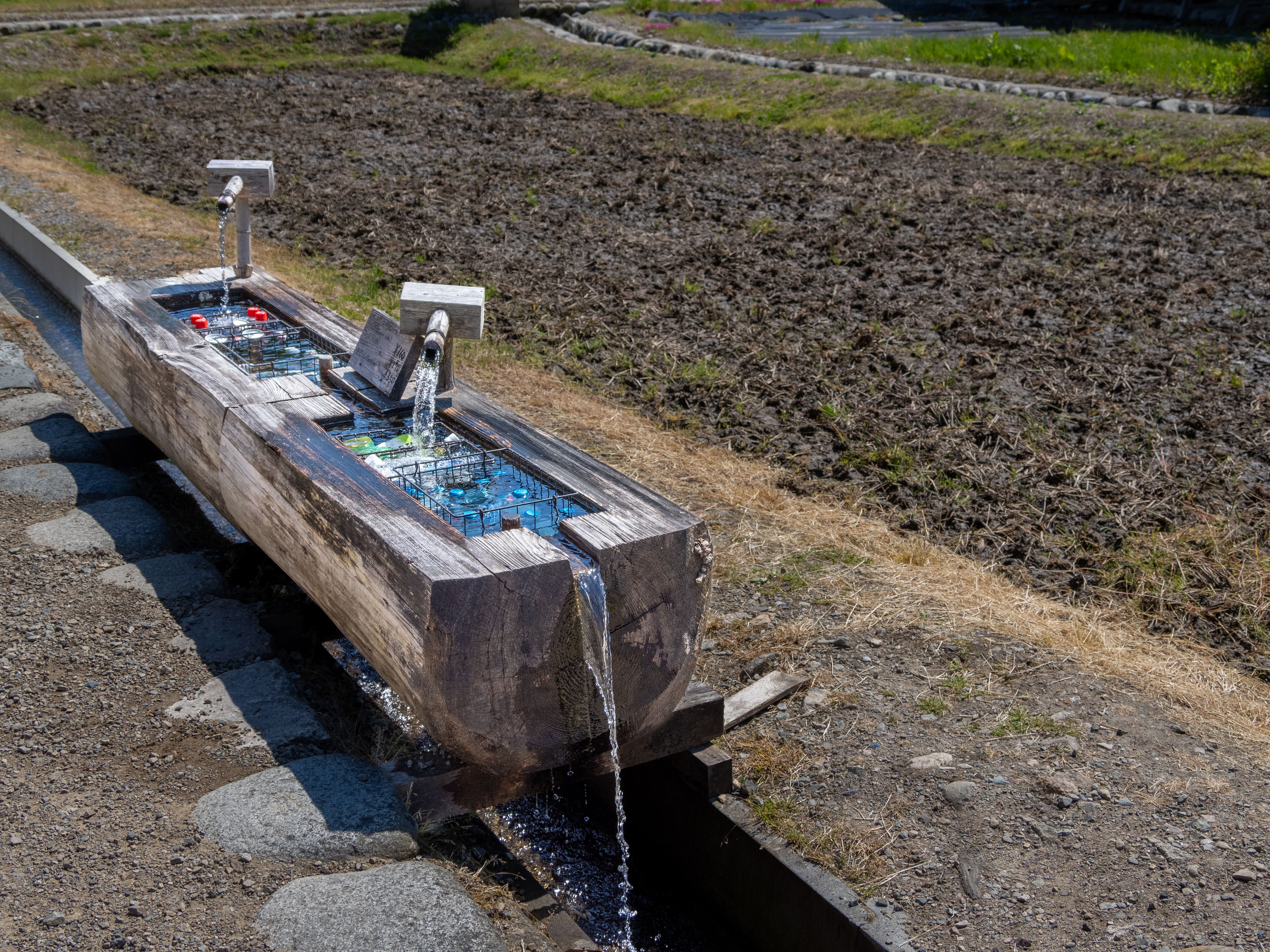 Wooden water trough in a Japanese village used for cooling drinks, with fields in the background.