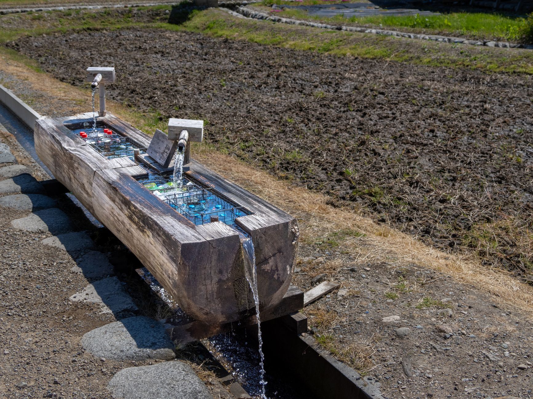 Wooden water trough in a Japanese village used for cooling drinks, with fields in the background.