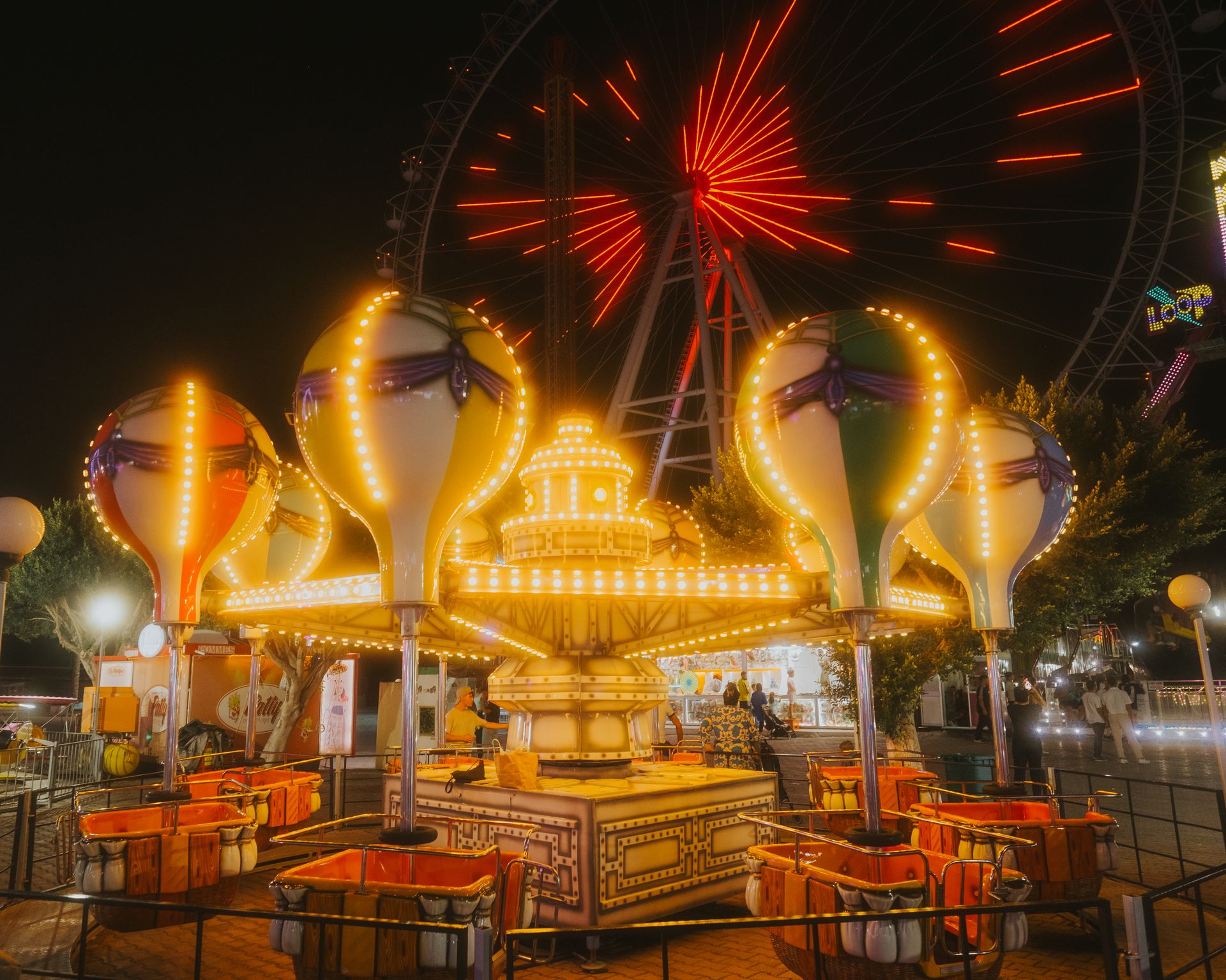 A vibrant night shot of a Ferris wheel and carousel at an amusement park in Antalya, Türkiye.