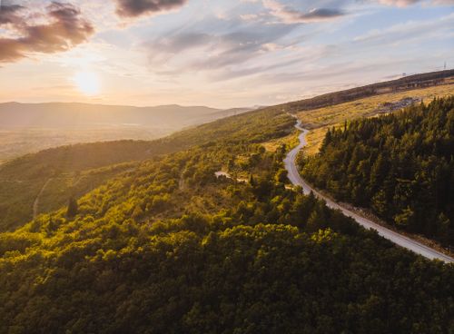 Free Stunning aerial view of a winding road through a lush green valley at sunset with a scenic mountain backdrop. Stock Photo