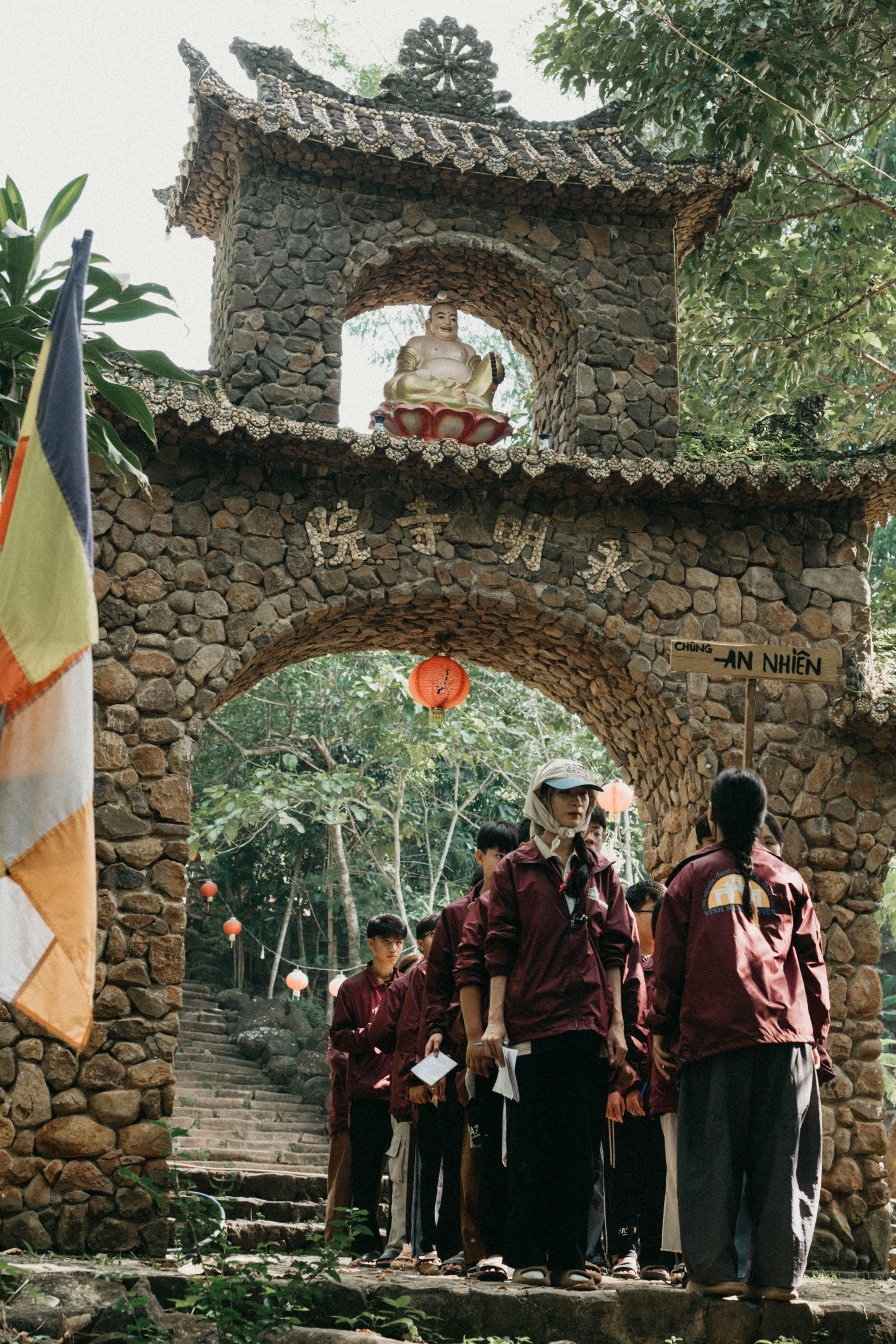 Visitors explore a stone temple gateway with a Buddha statue set in lush greenery.